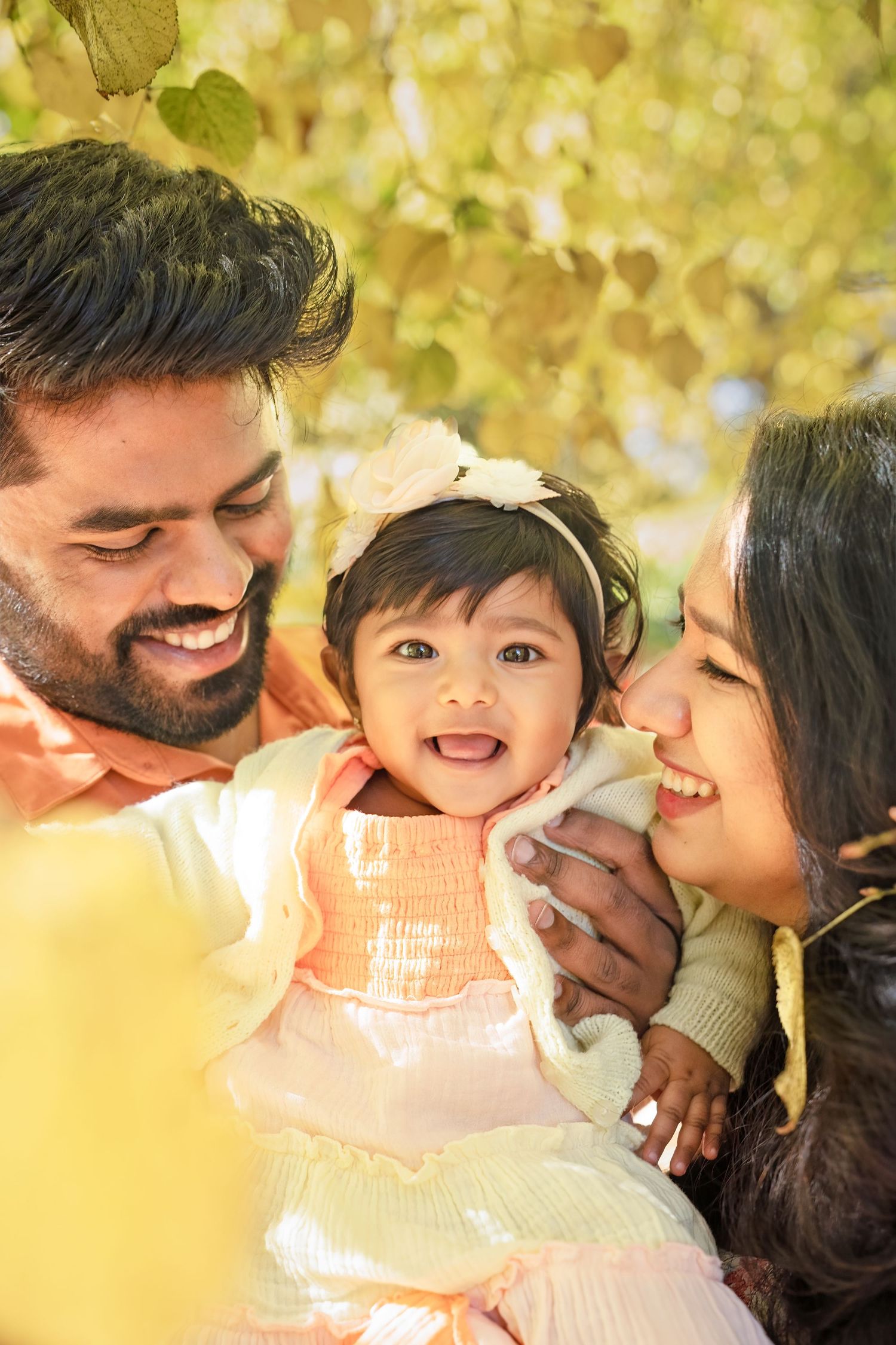Happy young family with smiling baby girl enjoying autumn park, parents kissing infant daughter outdoors.