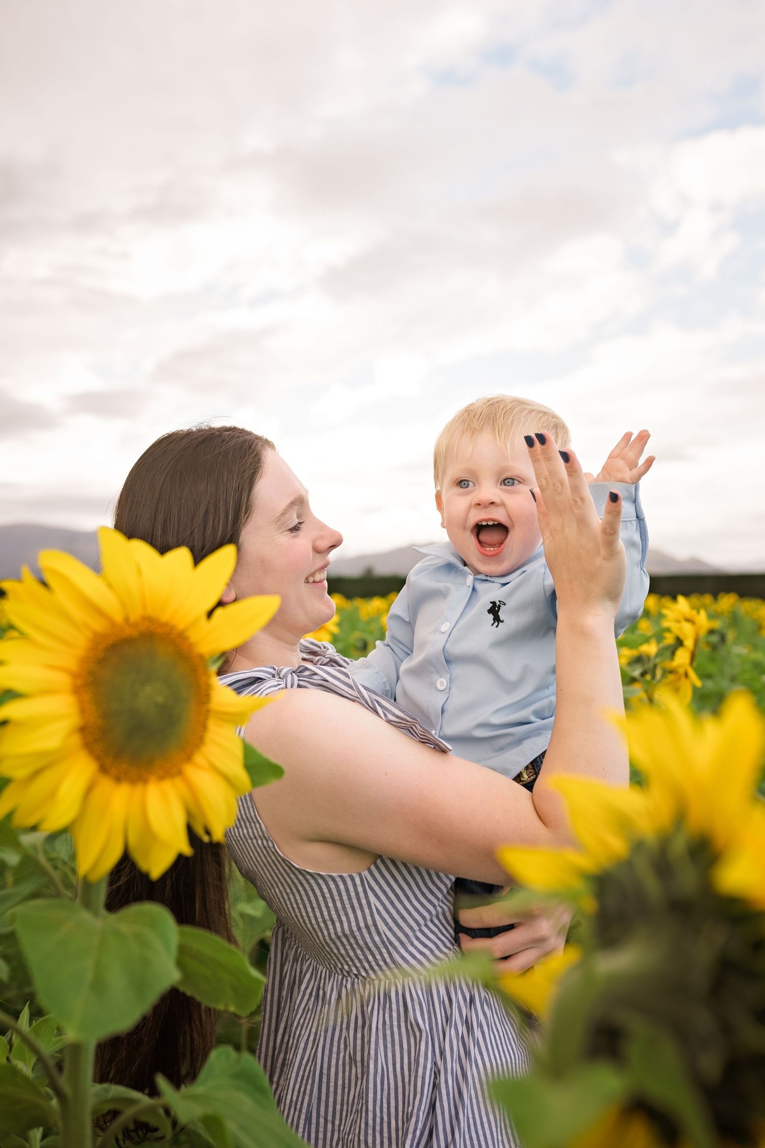 A smiling mother holds her laughing baby boy in a vibrant sunflower field under a cloudy sky.