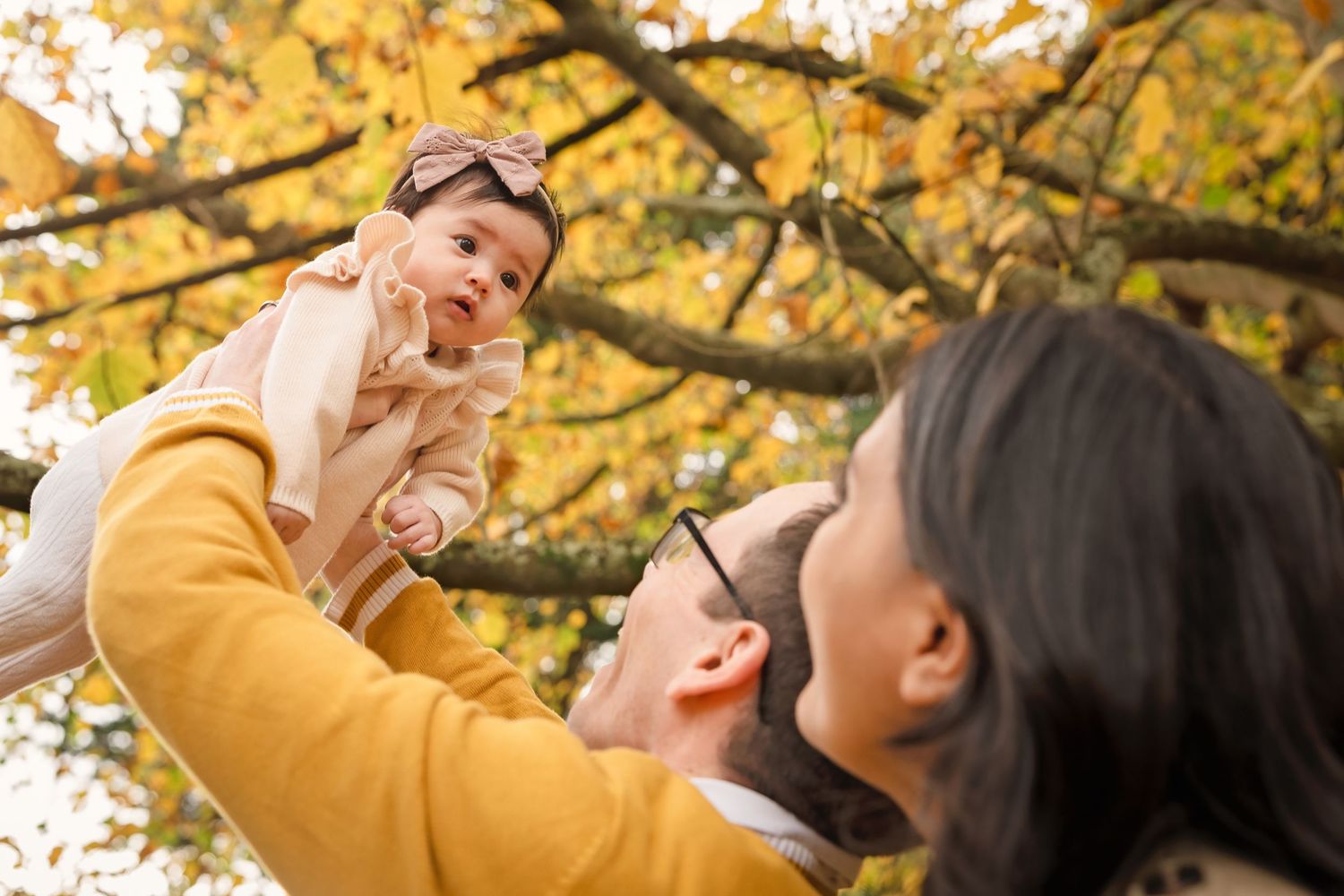 Parents lifting their baby in autumn park with golden leaves, family outdoor fall photoshoot.