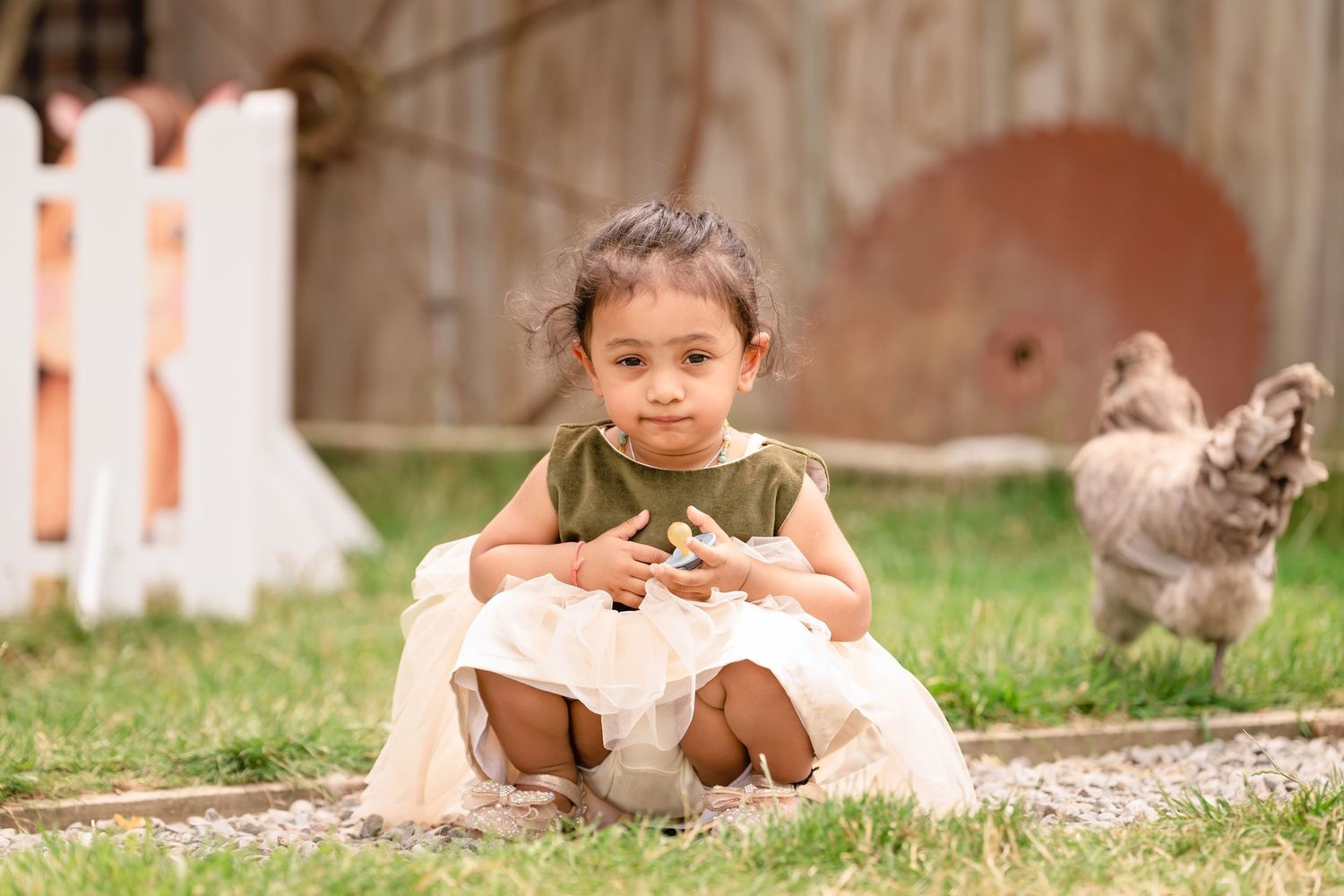 A young child in a dress sits outdoors on grass with chickens roaming in the background.