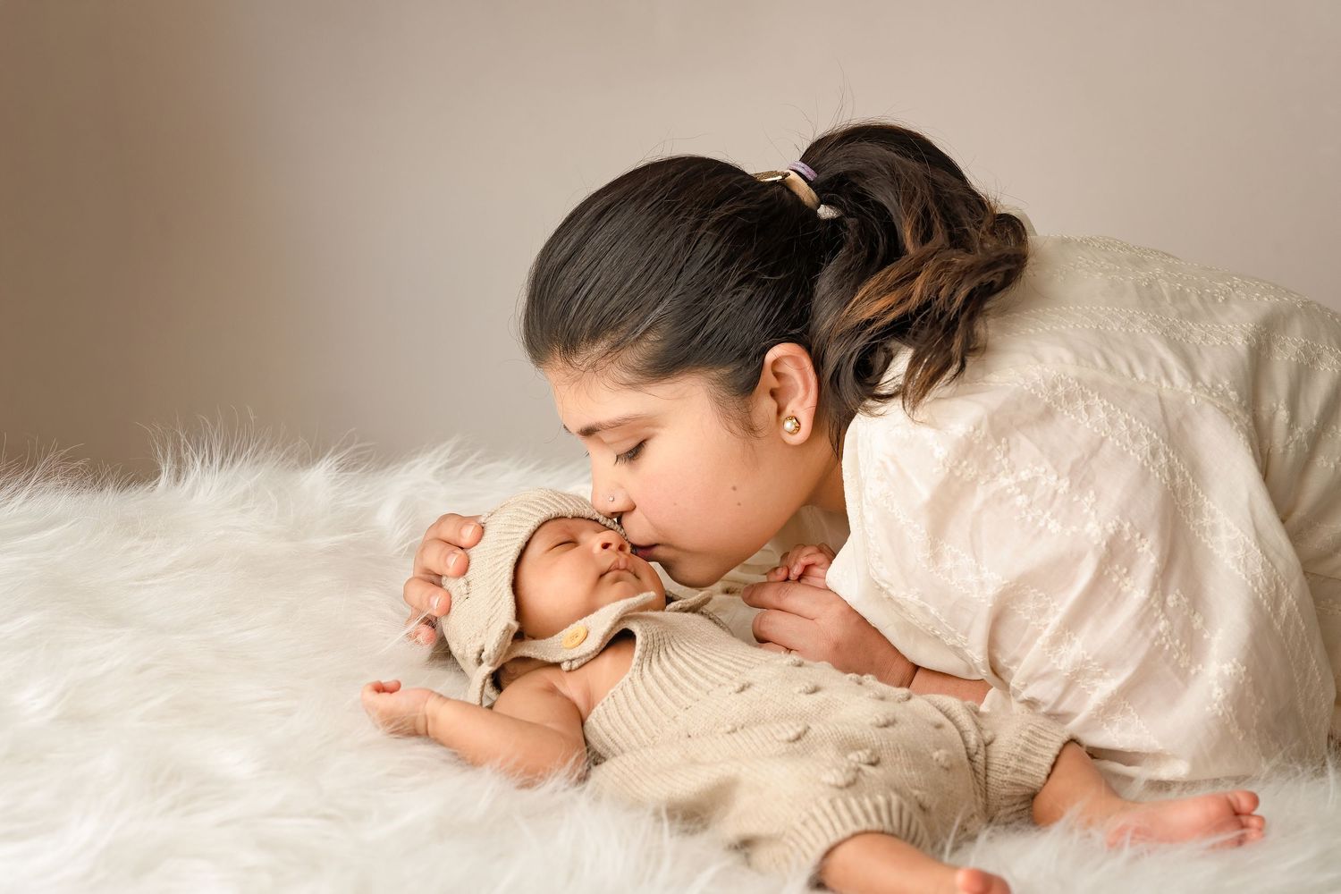 Older sister gently kissing newborn baby lying on white fluffy blanket in soft neutral tones.