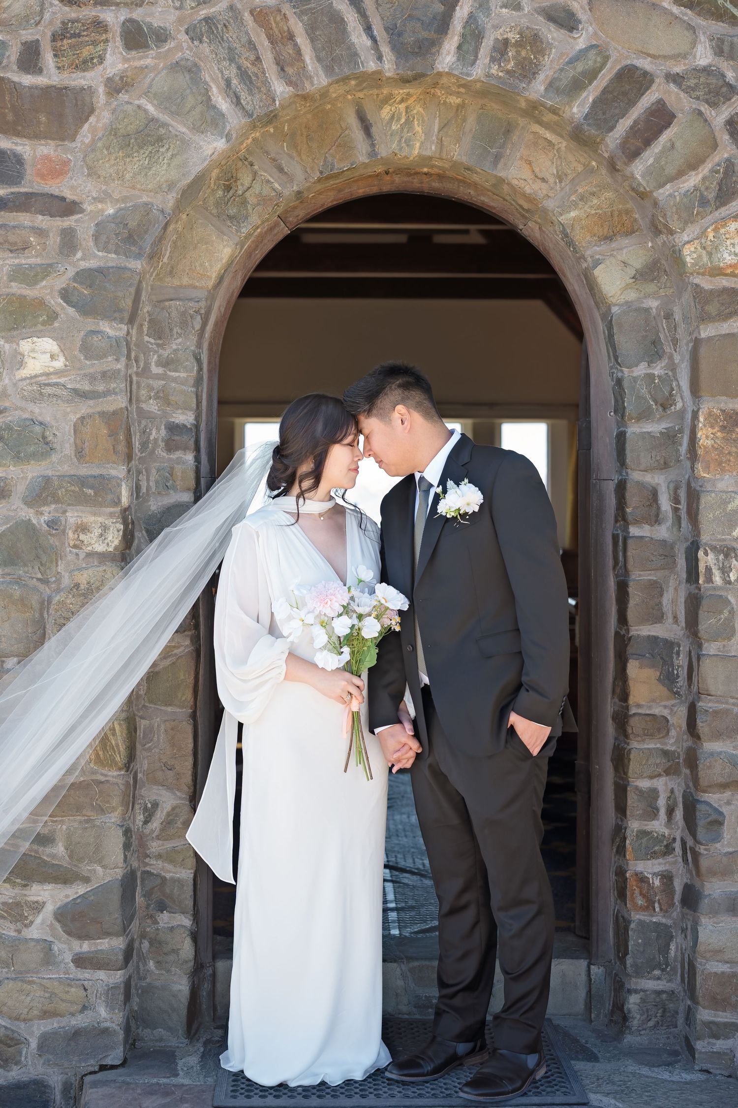 Bride and groom share an intimate moment in a stone church arched doorway, her veil flowing as she holds a white bouquet.