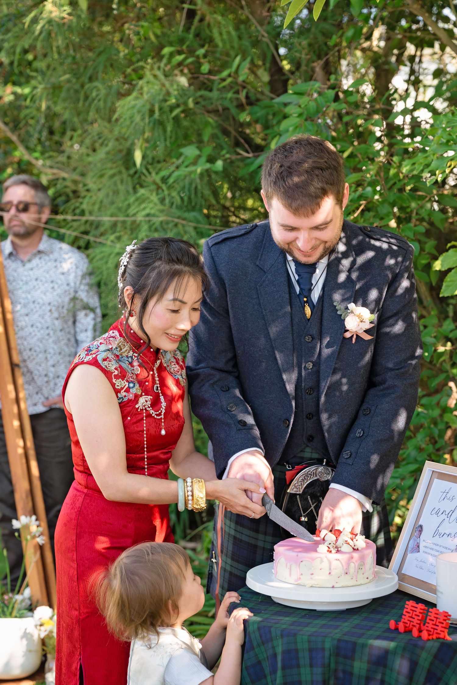 Bride in red qipao and groom in kilt cutting wedding cake outdoors at multicultural wedding celebration.