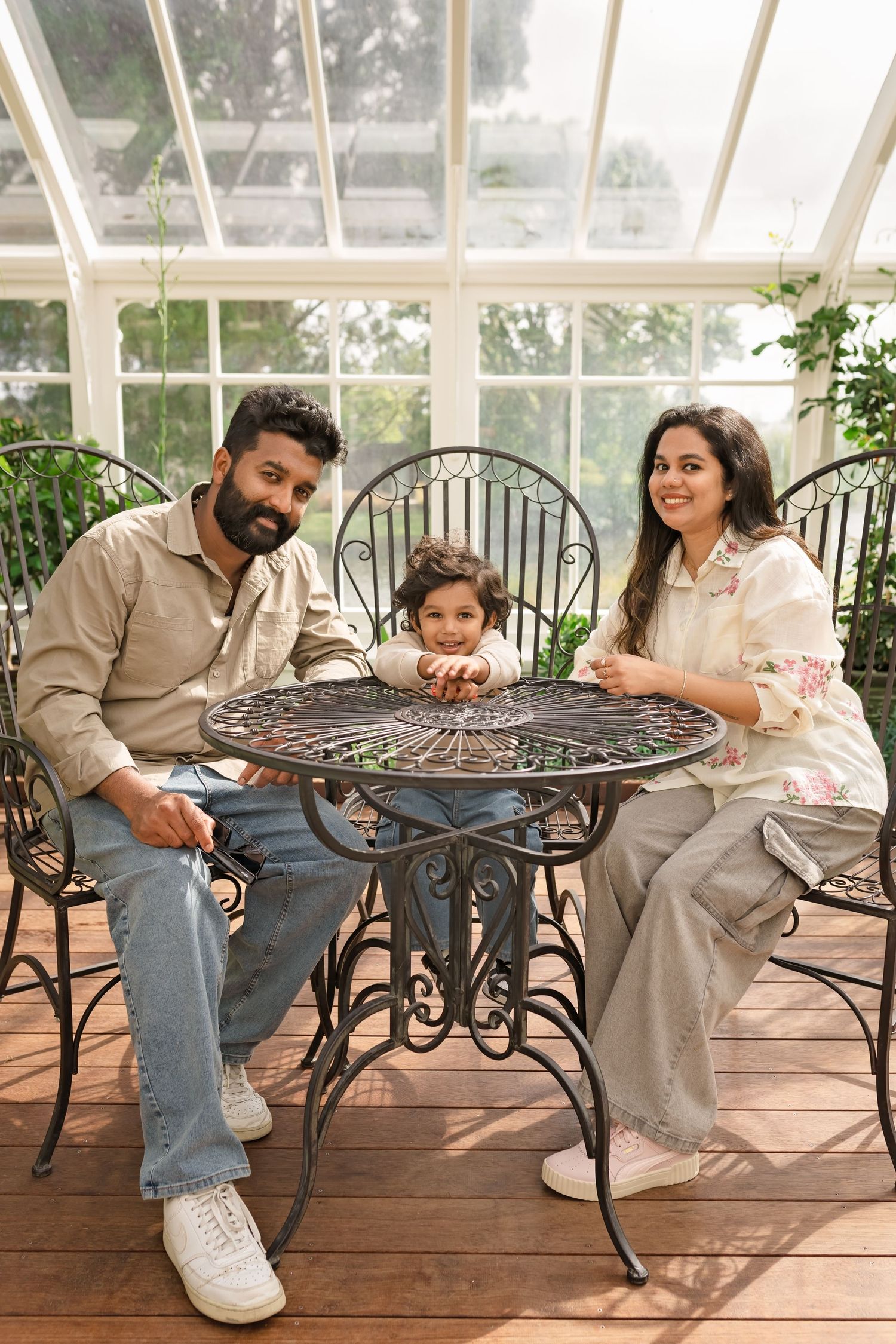 A happy South Asian family of three sits at an ornate iron table in a sunlit greenhouse conservatory.