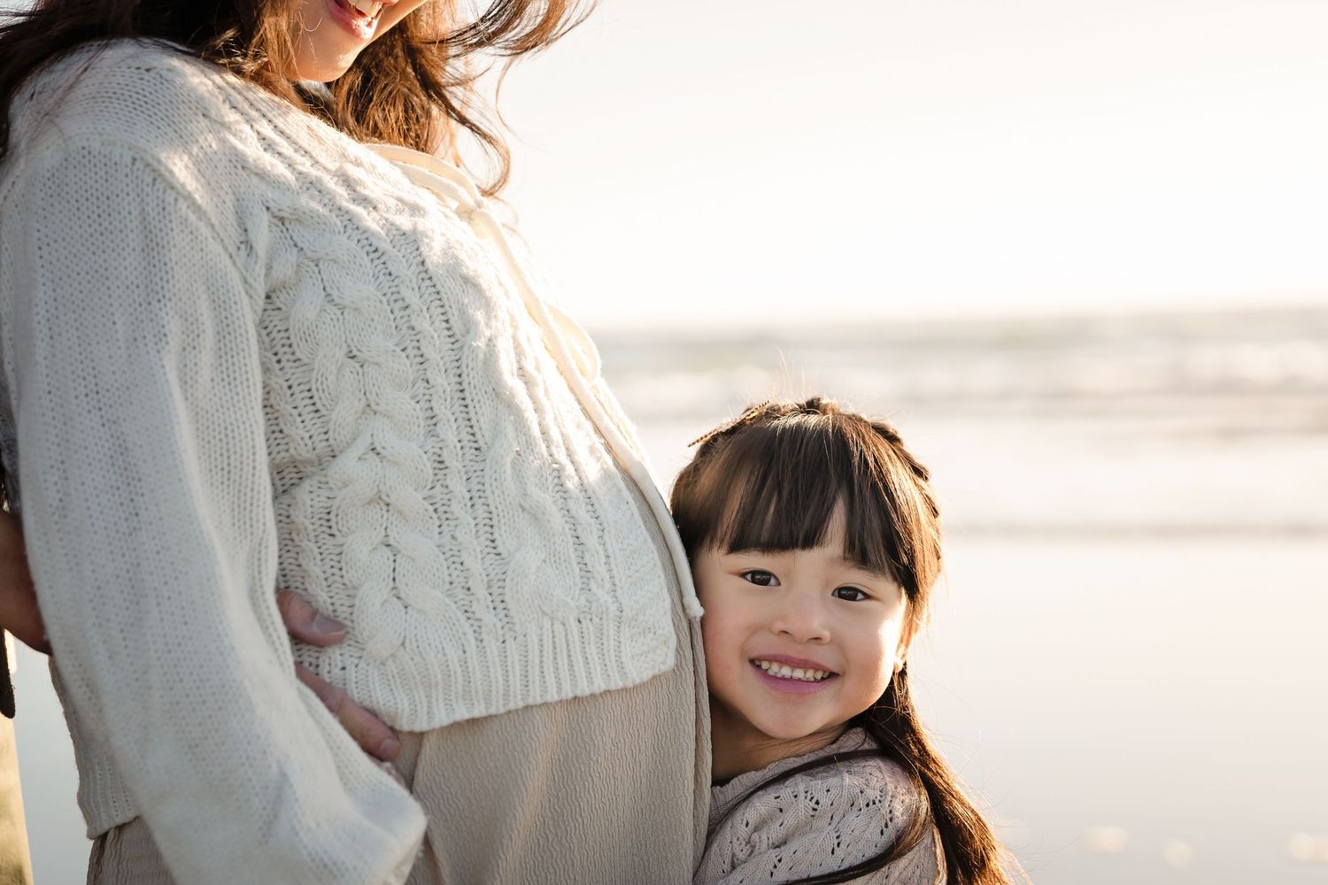 A young smiling Asian girl hugs her pregnant mother's belly on a beach, both wearing cozy cream sweaters.