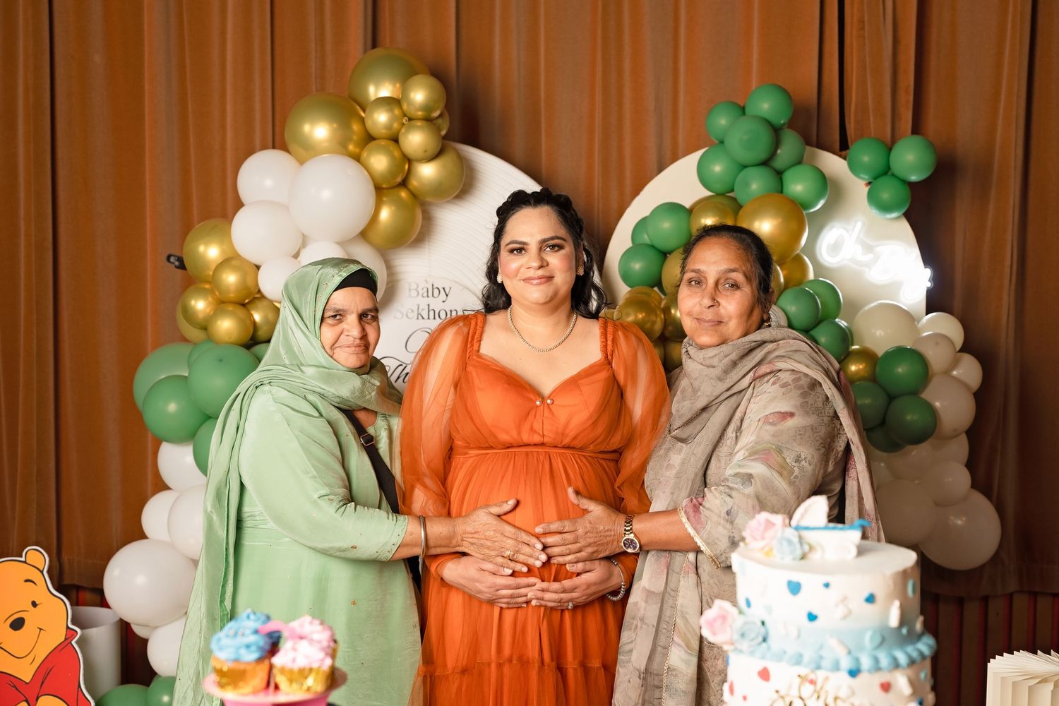 Three women pose together at a baby shower with green and white balloon arch, cake, and decorations.