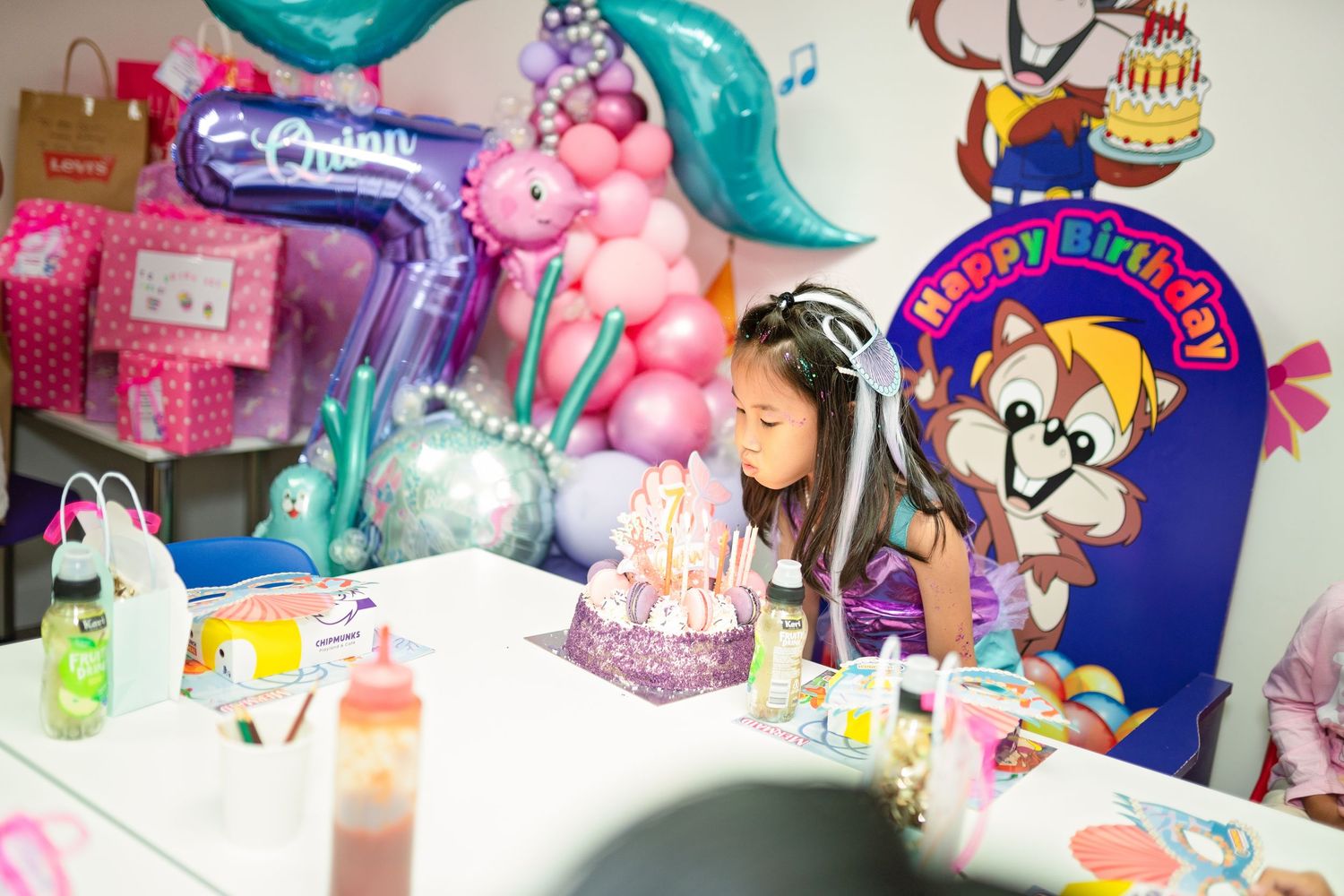 Young girl blowing out birthday cake candles at a mermaid-themed party with colorful balloon decorations.