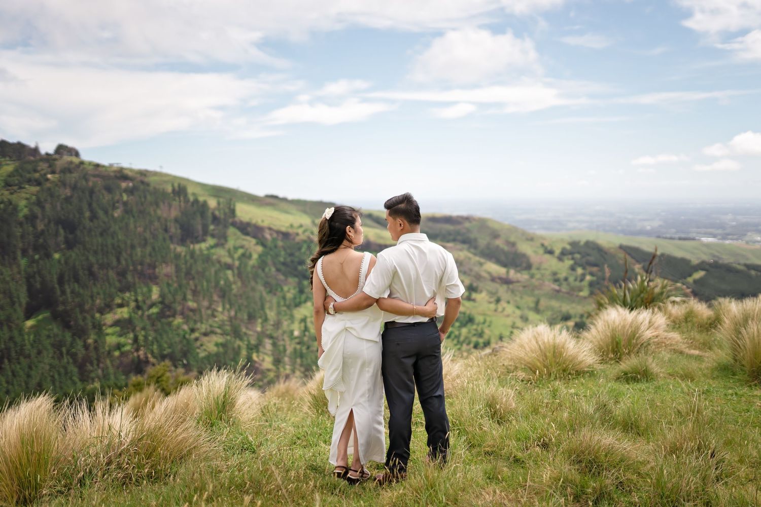 Couple embracing on a scenic hilltop with lush green valleys and dramatic cloudy skies in the background.
