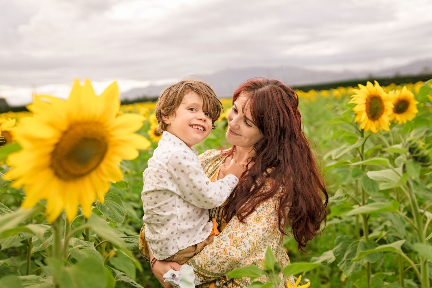 A smiling child and his mother share a joyful embrace surrounded by bright sunflowers.