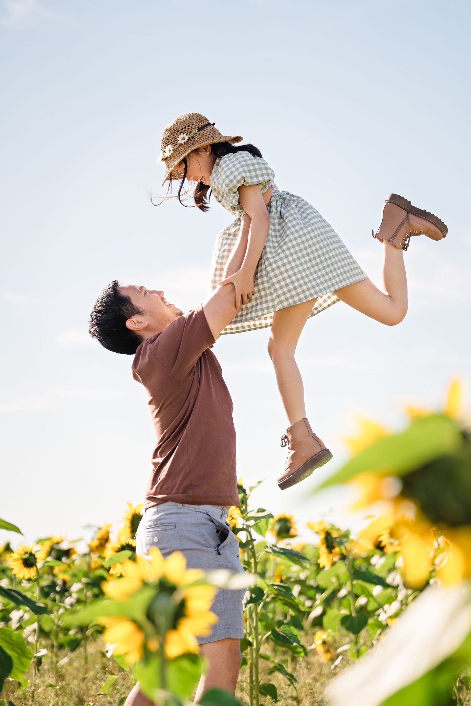 A person lifts a child playfully in the air above a field of blooming sunflowers on a bright sunny day.