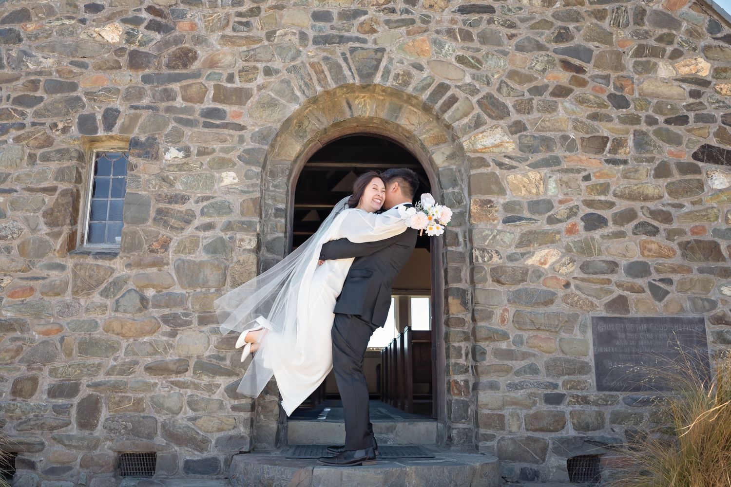 Groom lifting bride in wedding dress outside stone church archway.