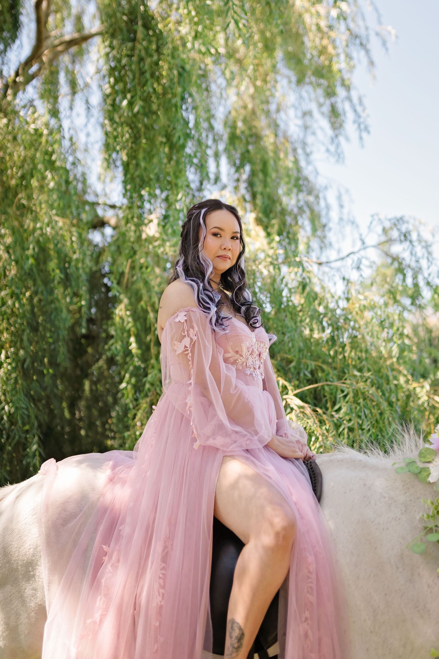 Woman in pink tulle gown sitting on white horse under weeping willow tree.