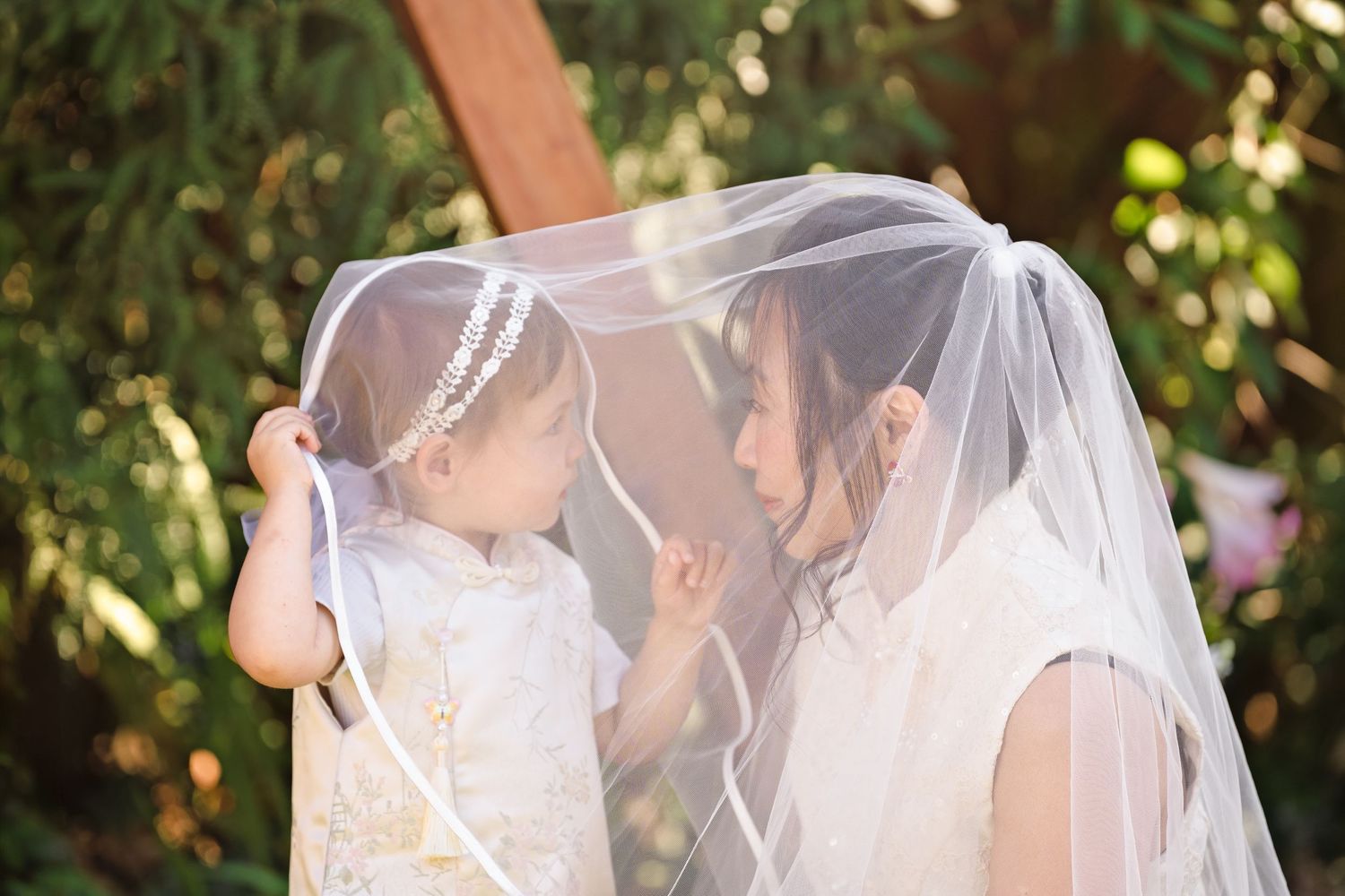 Bride and flower girl sharing a tender moment under a white wedding veil outdoors.