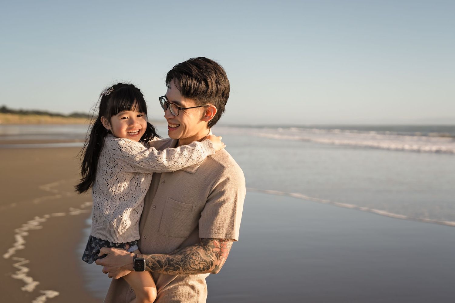 Father holding smiling daughter on sunny beach, ocean waves in background.