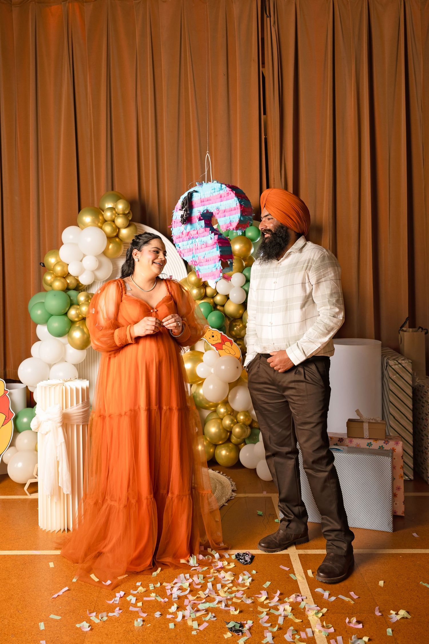 Couple at baby shower with colorful balloon arch and pinata, woman in orange maternity dress smiling.