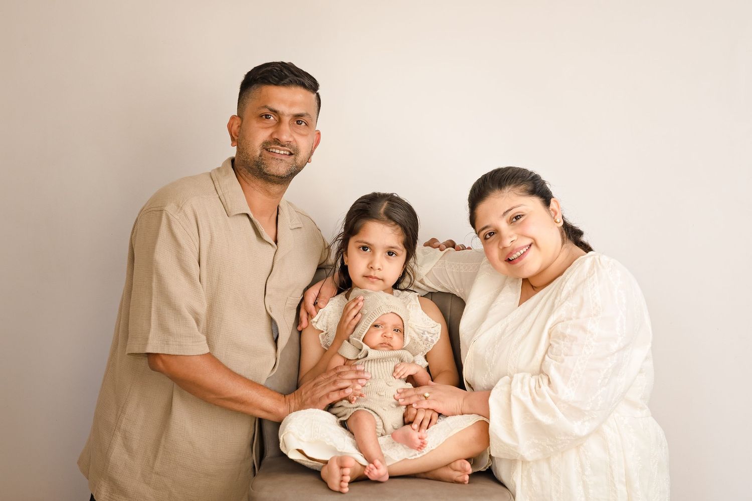 Family of four posing for portrait, parents with toddler girl holding newborn baby, neutral tones, studio setting.