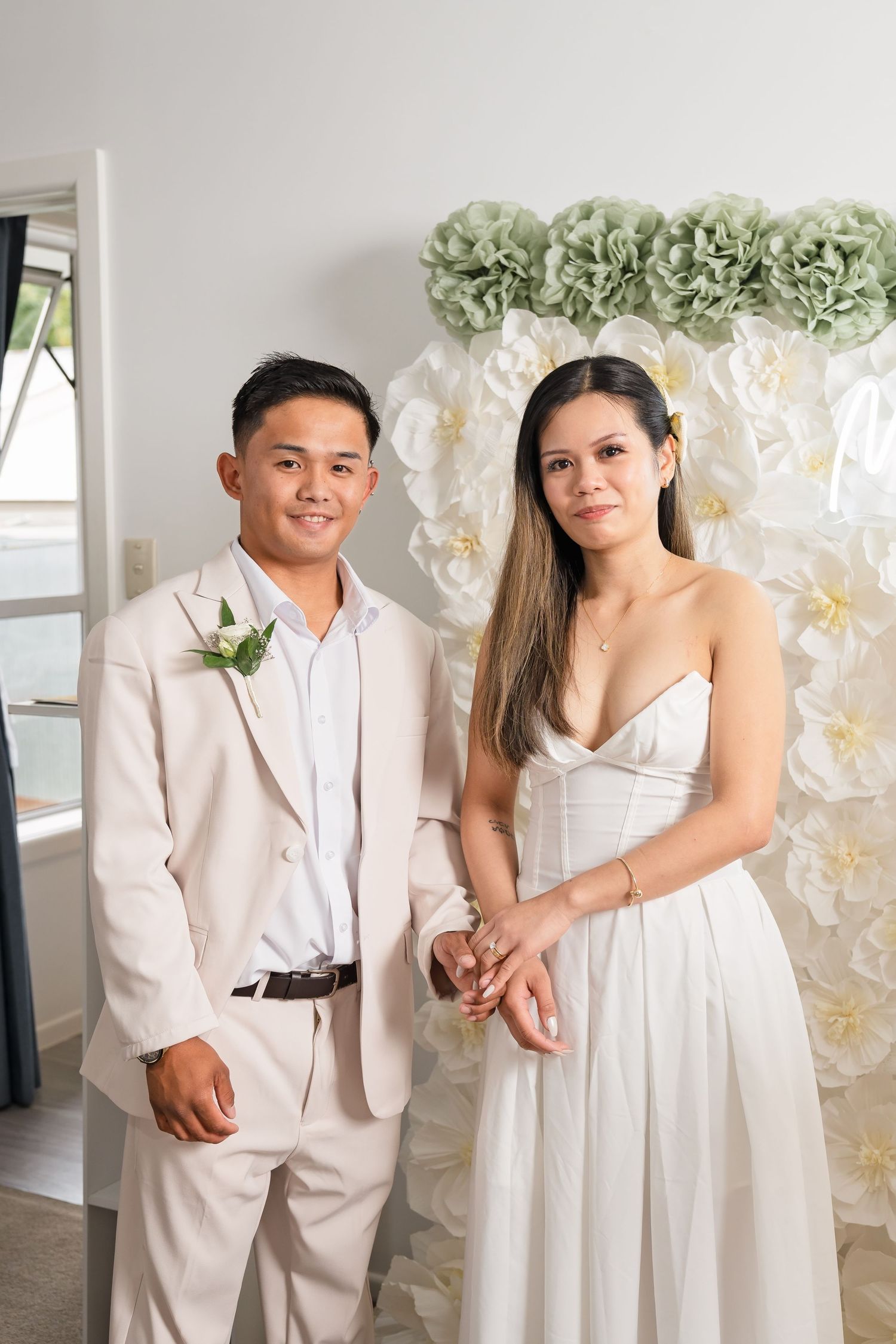 A couple in wedding attire poses before a floral backdrop, the groom in a beige suit and bride in a white gown.