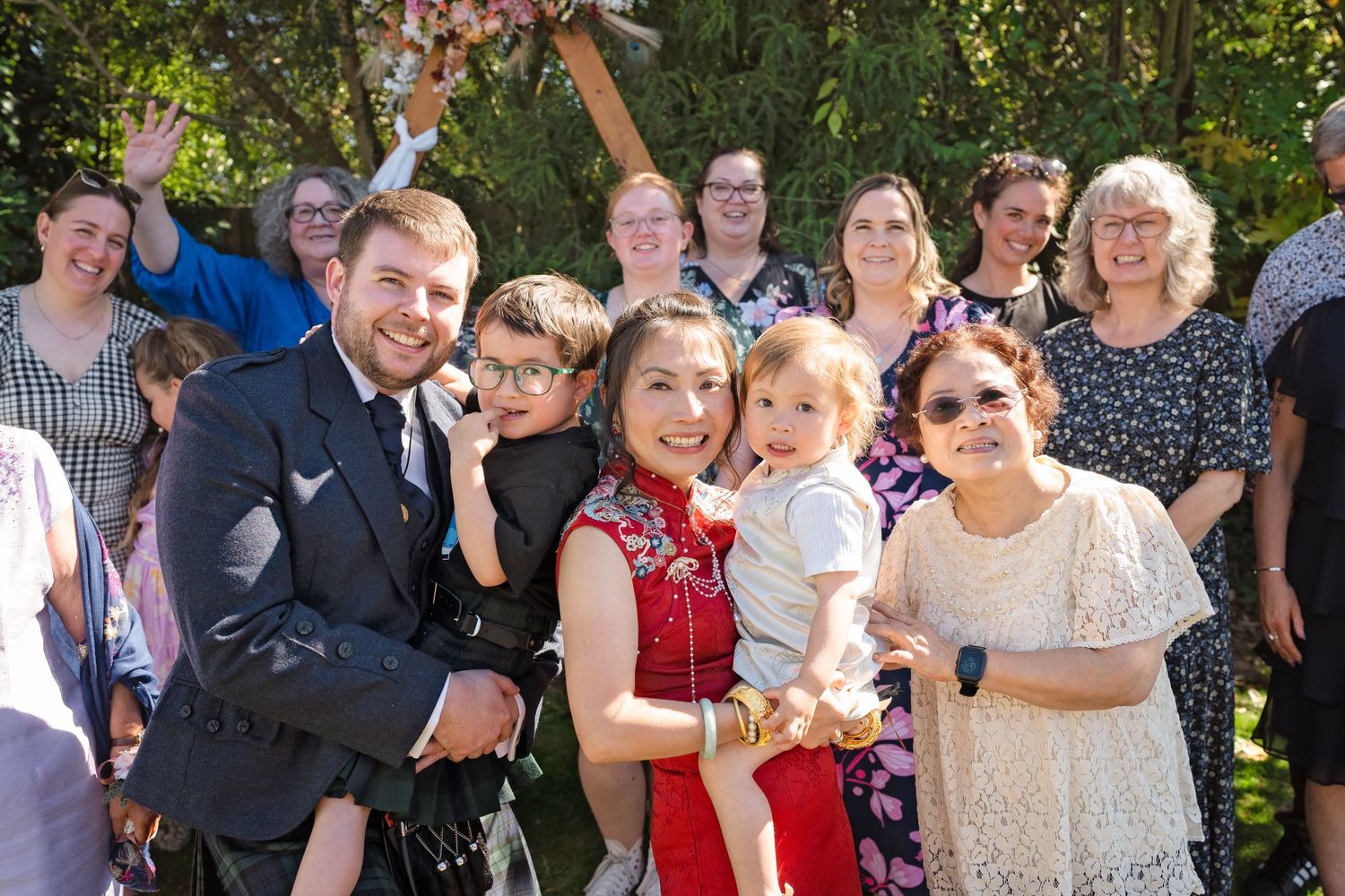Joyful wedding group photo with groom in suit, guests in colorful attire, and a baby outdoors.