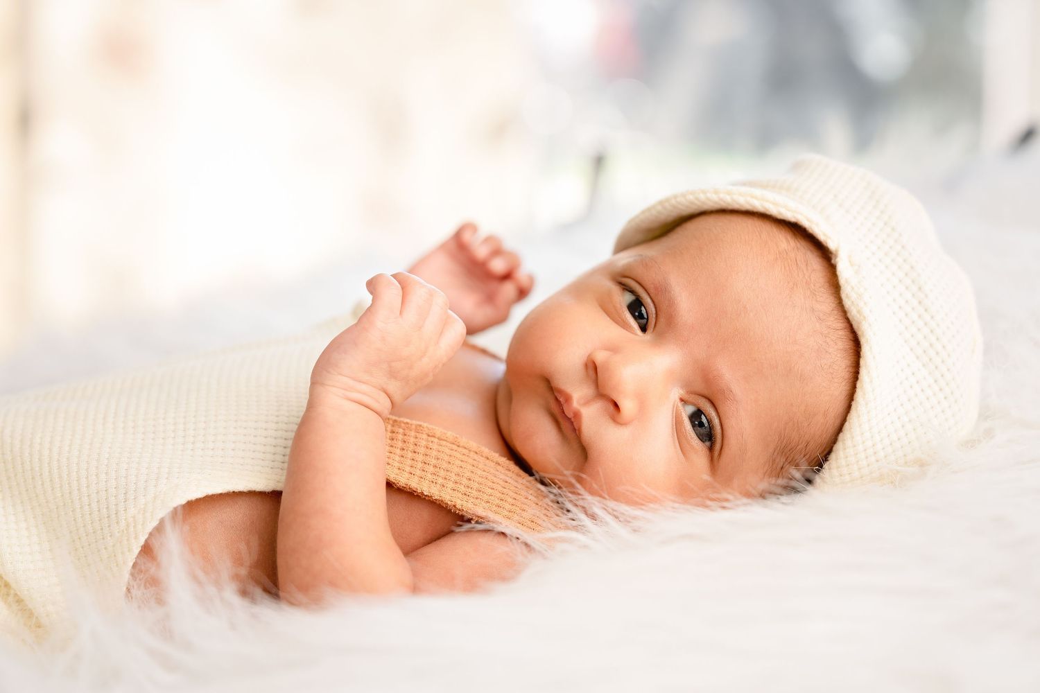 Newborn baby wearing a white knit hat lying on a fluffy white blanket, looking at the camera with bright eyes.