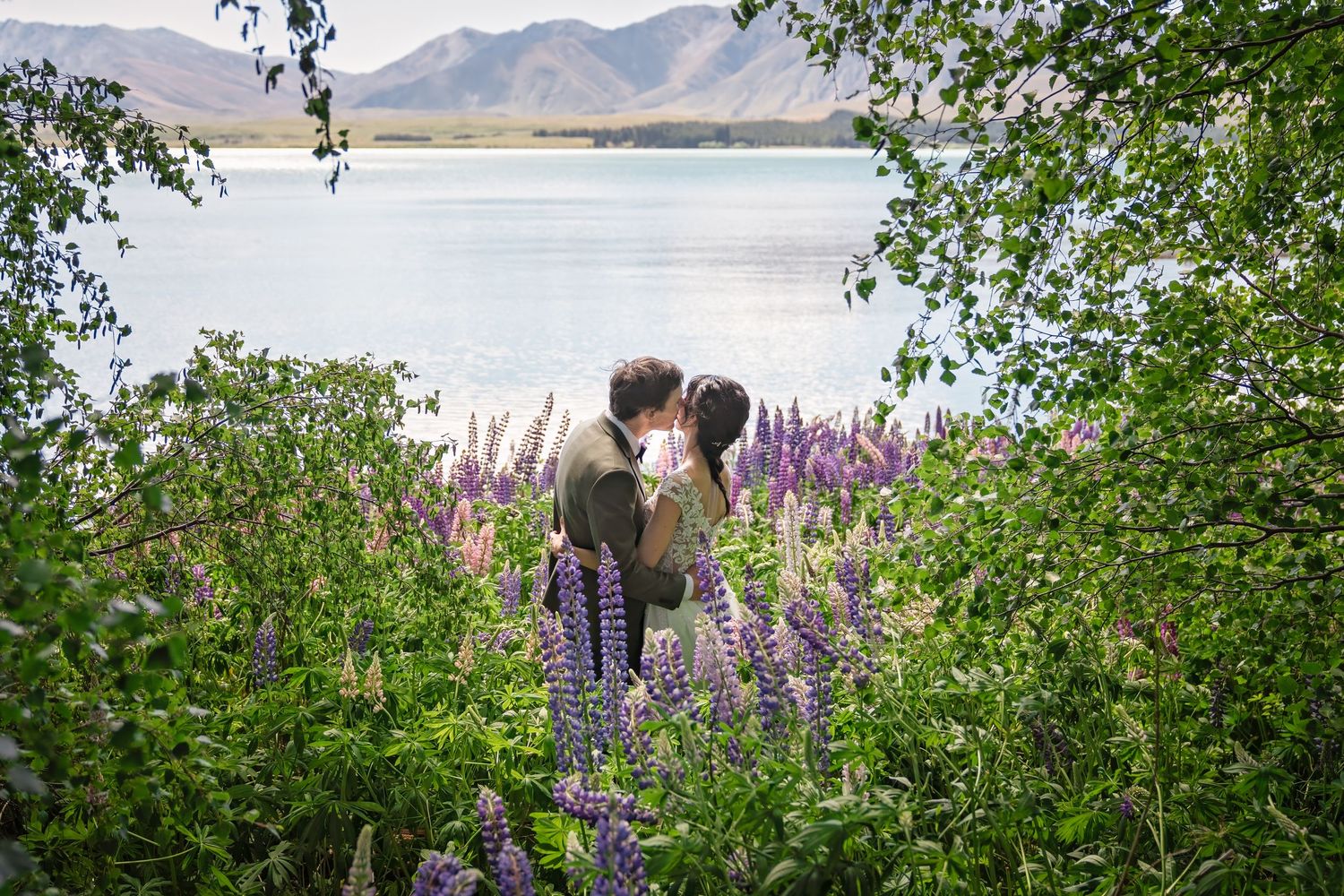 Two people stand among purple wildflowers beside a lake with mountains in the background.