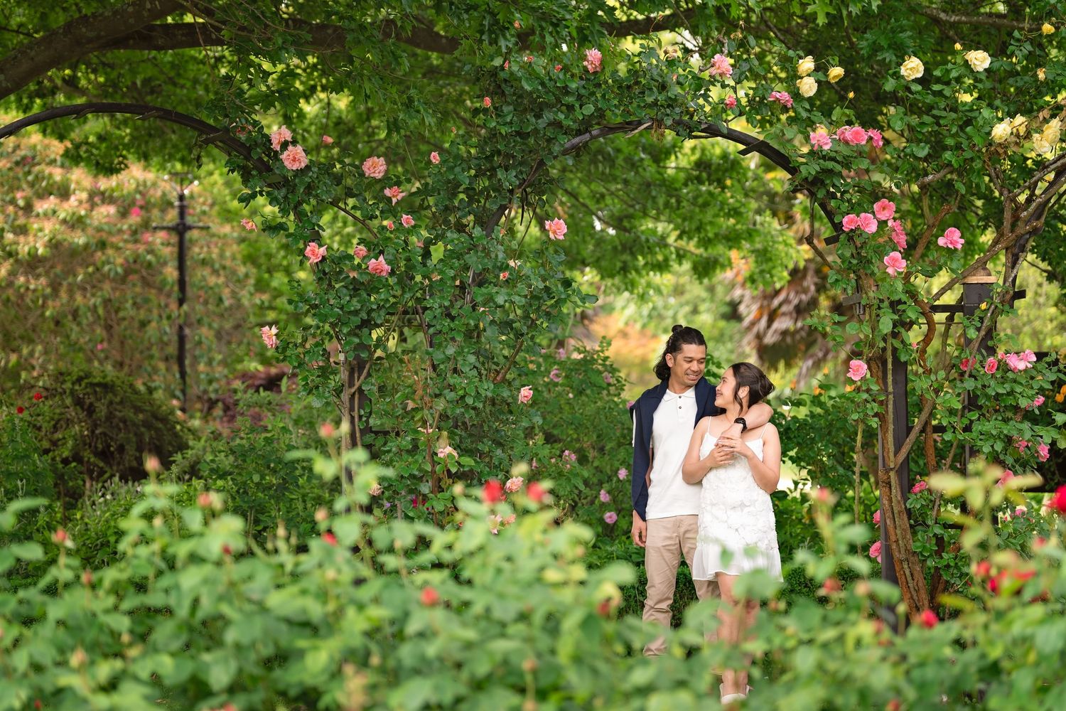 Couple sharing a romantic moment under a rose-covered garden arch surrounded by lush greenery.