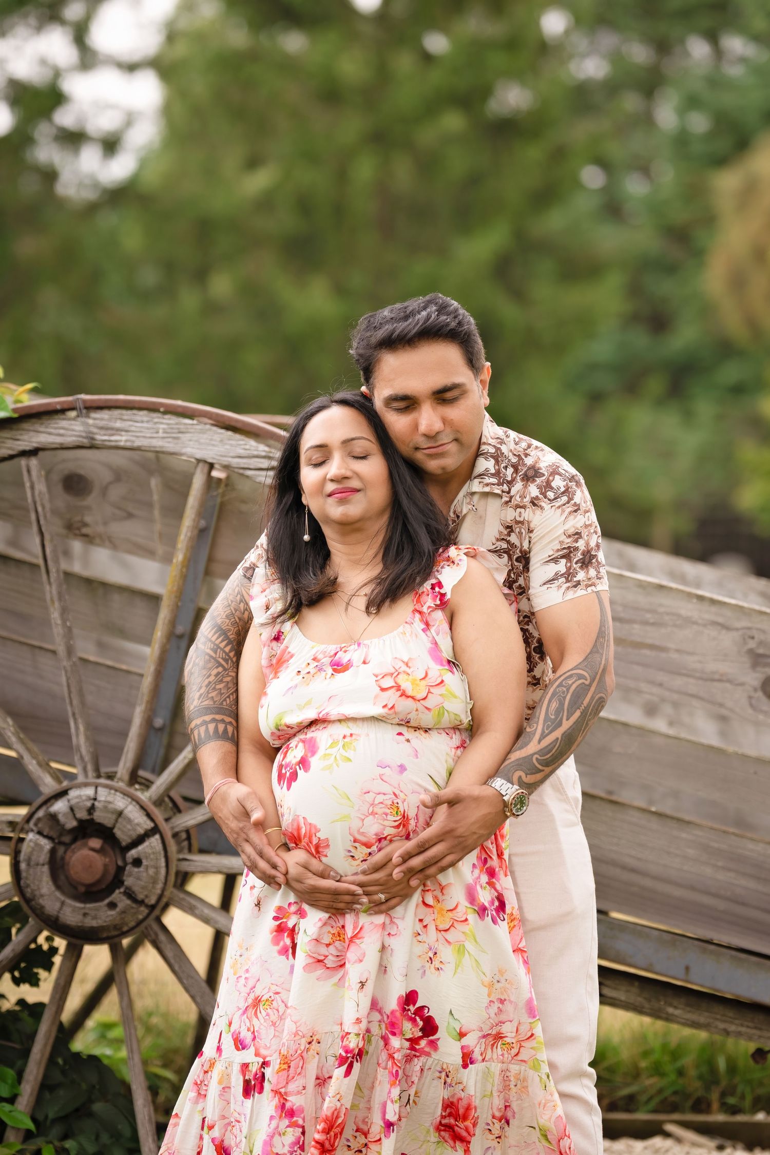 A couple in white and floral attire poses together against a rustic wooden backdrop during a maternity shoot.