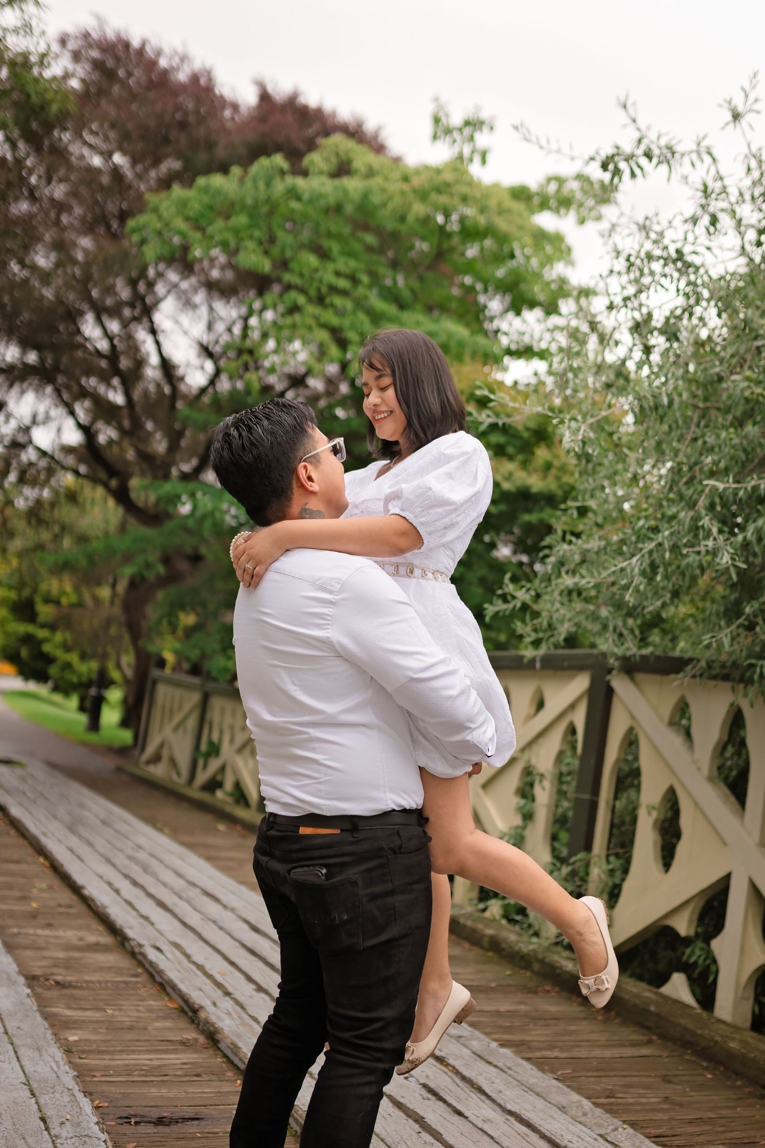 A man in white shirt lifts a smiling woman on a wooden bridge surrounded by lush green trees.