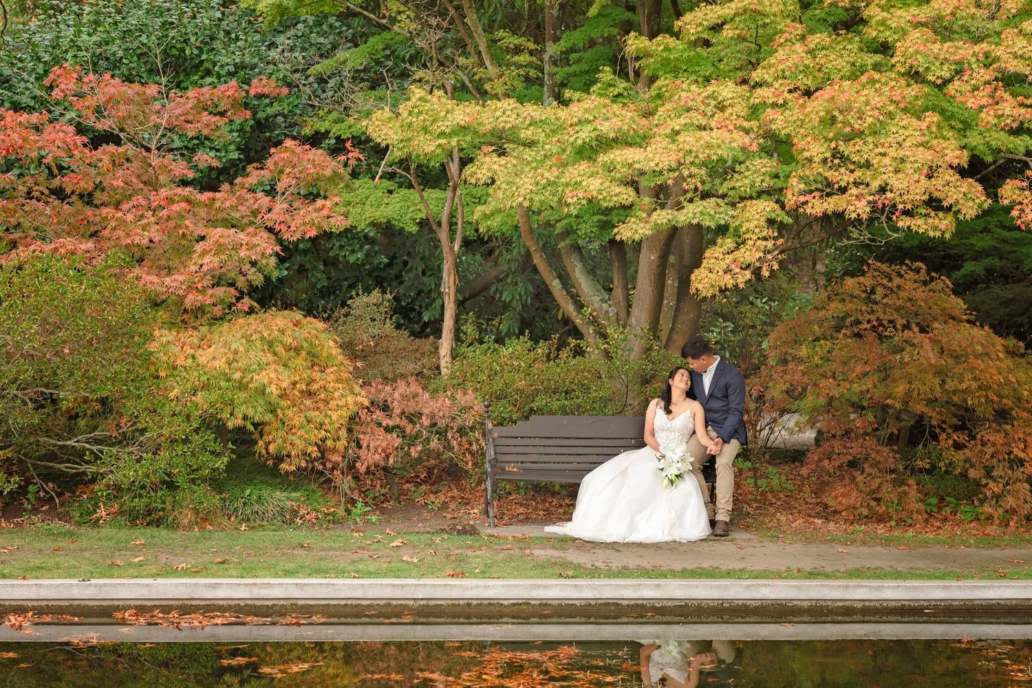 Bride and groom sitting on a bench by a pond surrounded by colorful autumn foliage in a serene garden.
