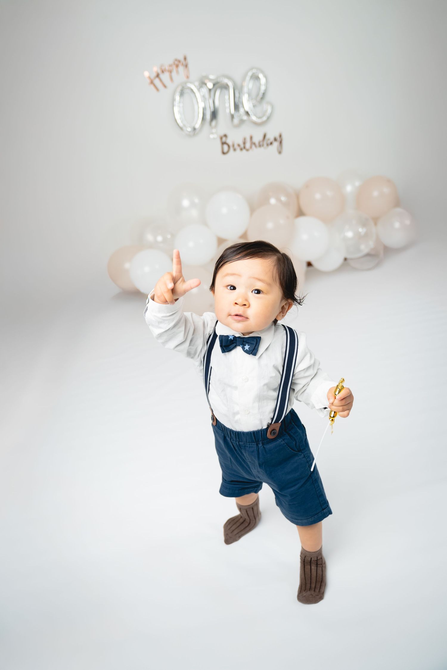 A toddler in navy suspenders and bow tie poses under silver 'ONE' birthday balloons.