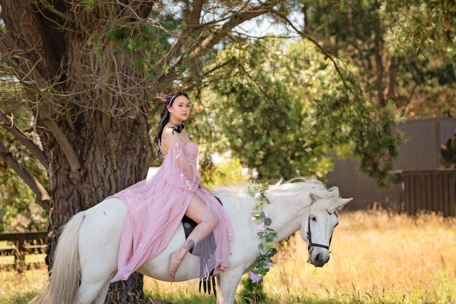Young woman in pink gown riding a white horse adorned with flowers beneath large trees in a sunlit field.