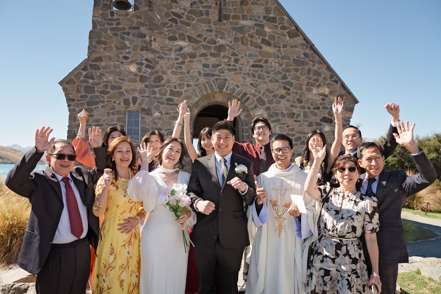 Asian wedding party celebrating outside a stone church with mountains in background.