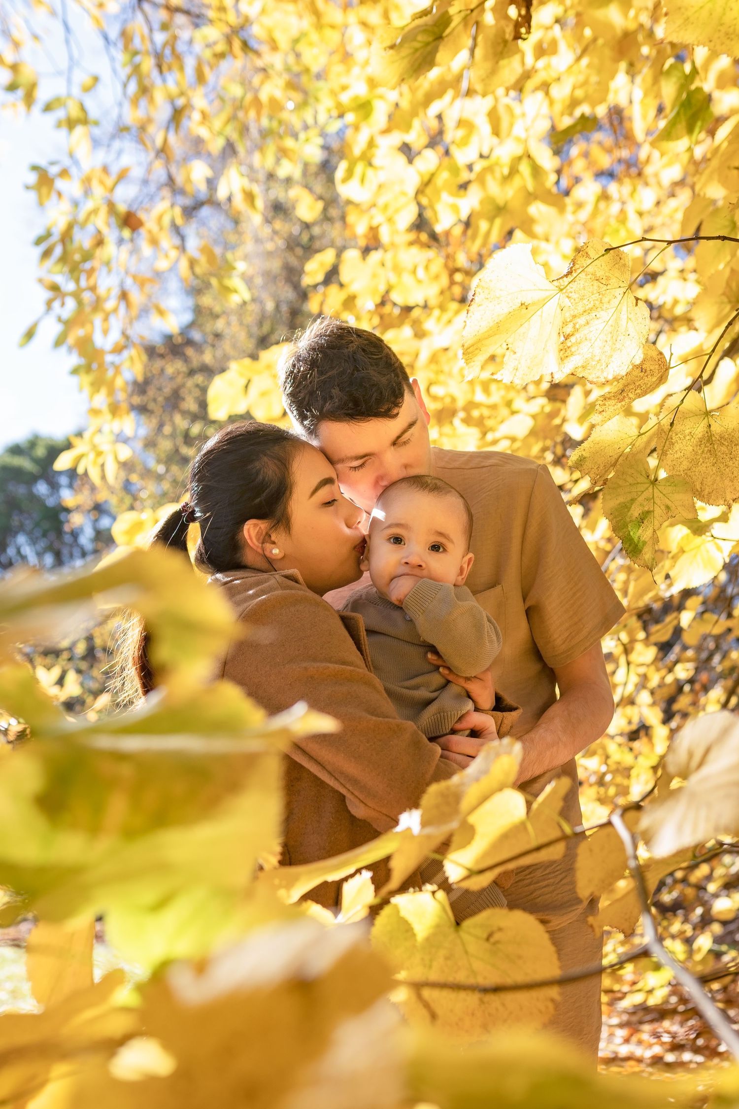 A tender family moment under golden autumn leaves as parents embrace their baby in matching brown outfits.