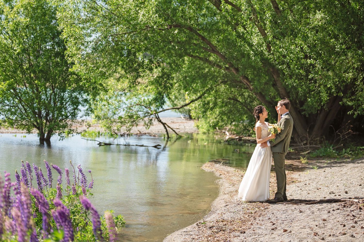 A couple in wedding attire poses along a scenic riverbank with purple wildflowers and trees in the background.