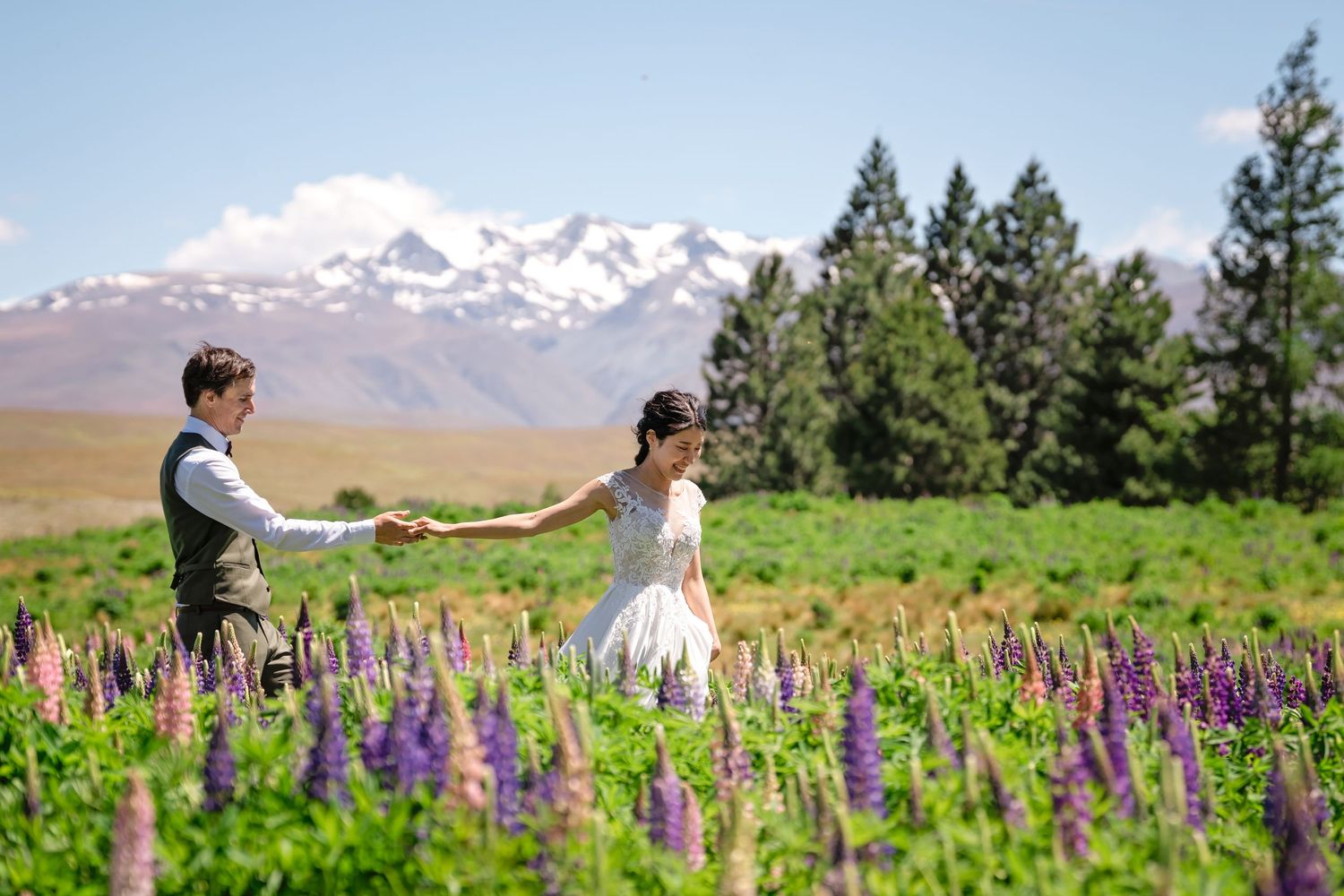 A couple walks through a vibrant field of purple lupine wildflowers with snow-capped mountains in the background.