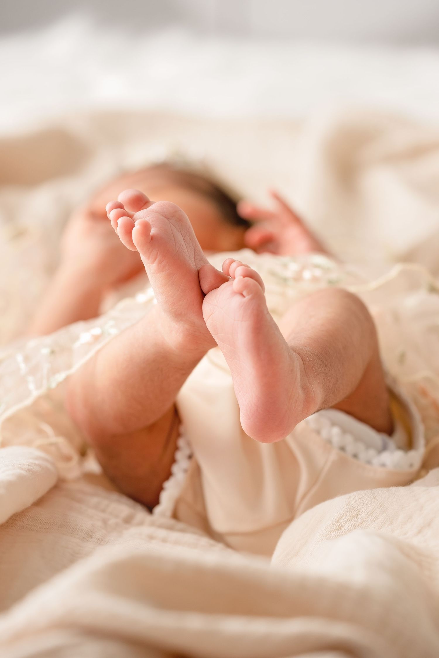 Close-up of tiny newborn baby feet in a cream lace outfit, lying on a soft beige blanket.