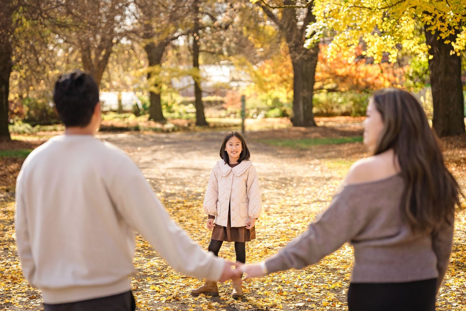 Family of three holding hands in a golden autumn park, parents facing daughter standing among fallen leaves.
