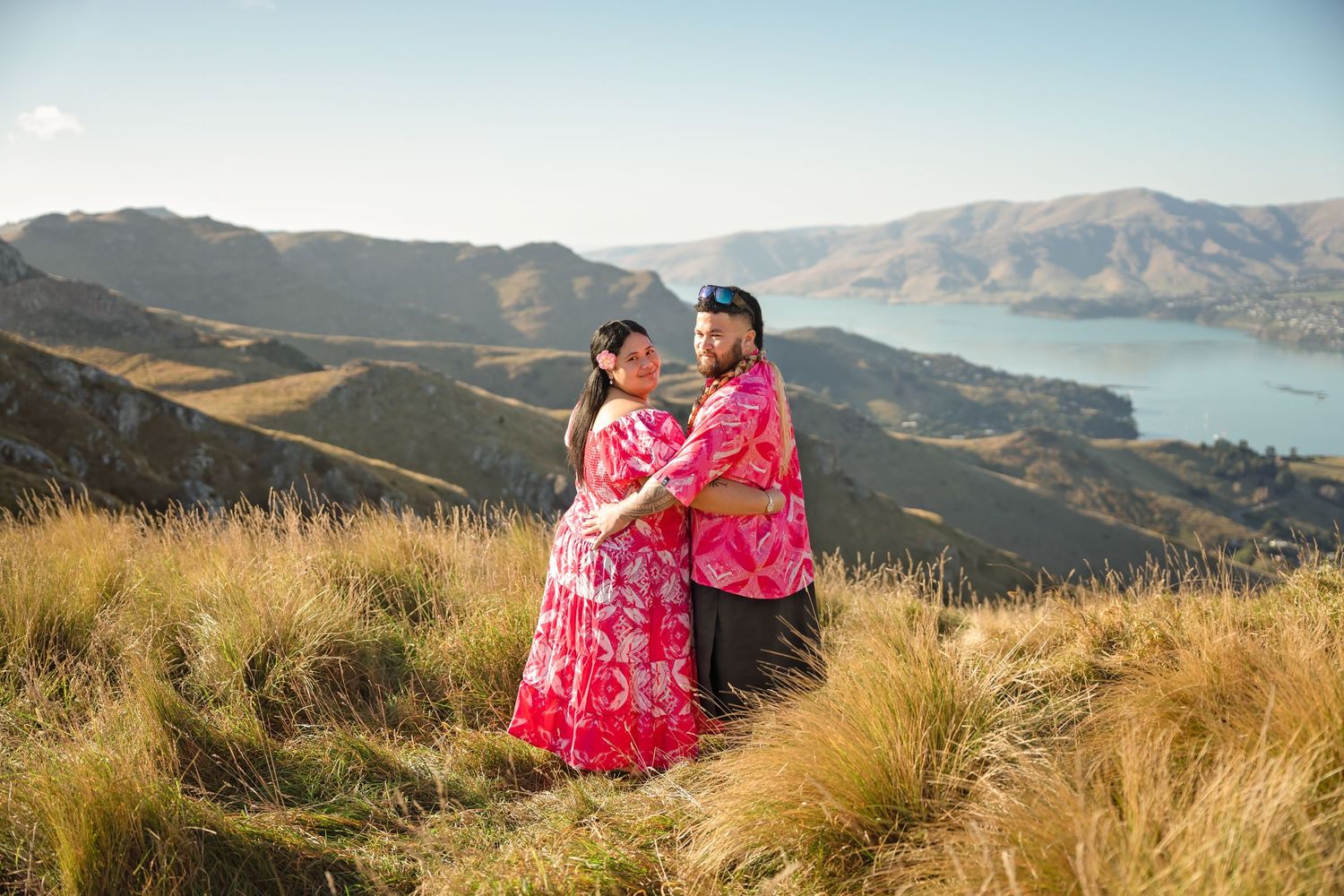 Couple in vibrant pink outfits embracing on a mountain peak with stunning lake and valley views in New Zealand.