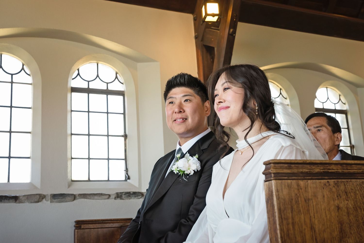 Couple smiling together in a chapel during a wedding ceremony, groom in dark suit with white boutonniere.