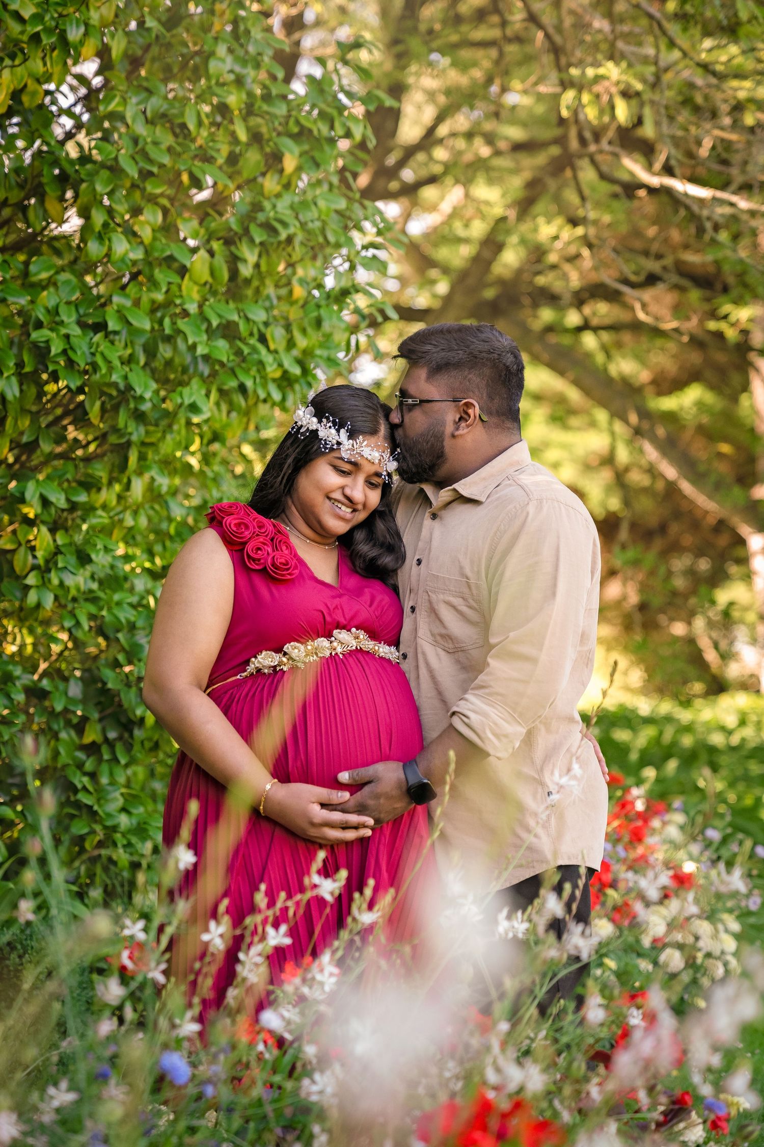 Expecting couple in garden, woman in red maternity gown with floral crown, man in beige shirt, surrounded by flowers.
