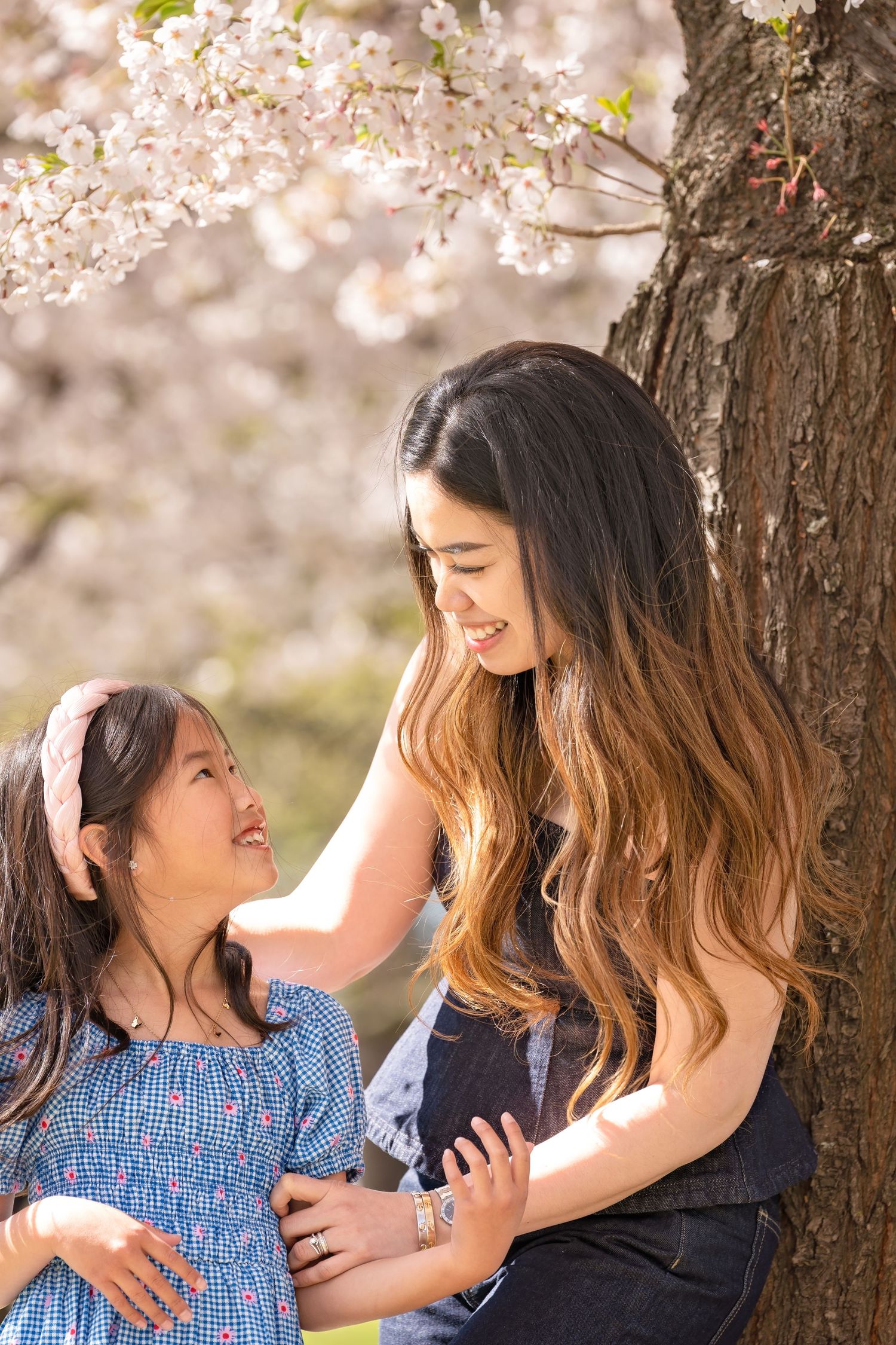 A woman and young girl smile at each other under blooming cherry blossom trees in spring.