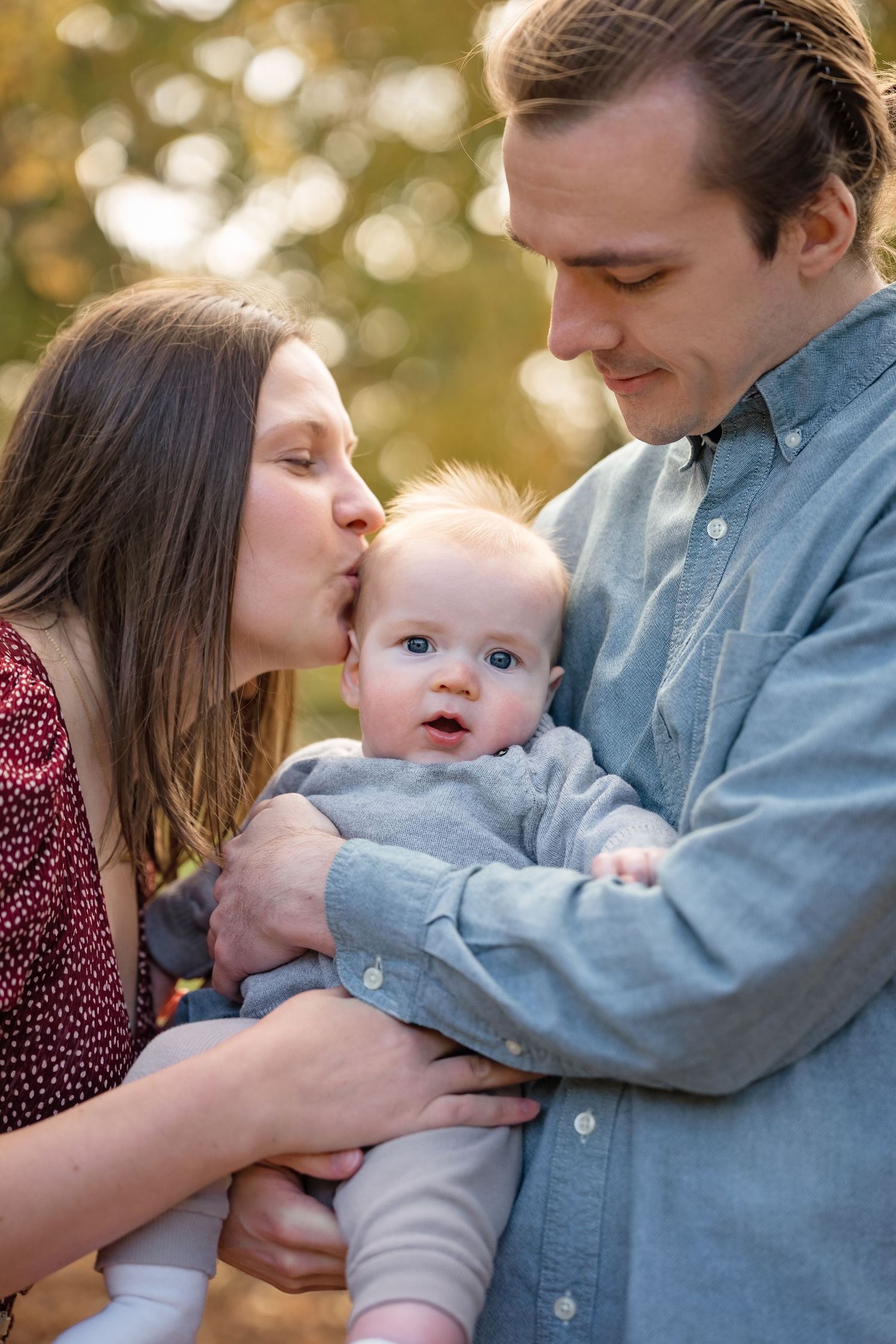 A young couple shares a tender moment kissing their blue-eyed baby outdoors in warm golden light.