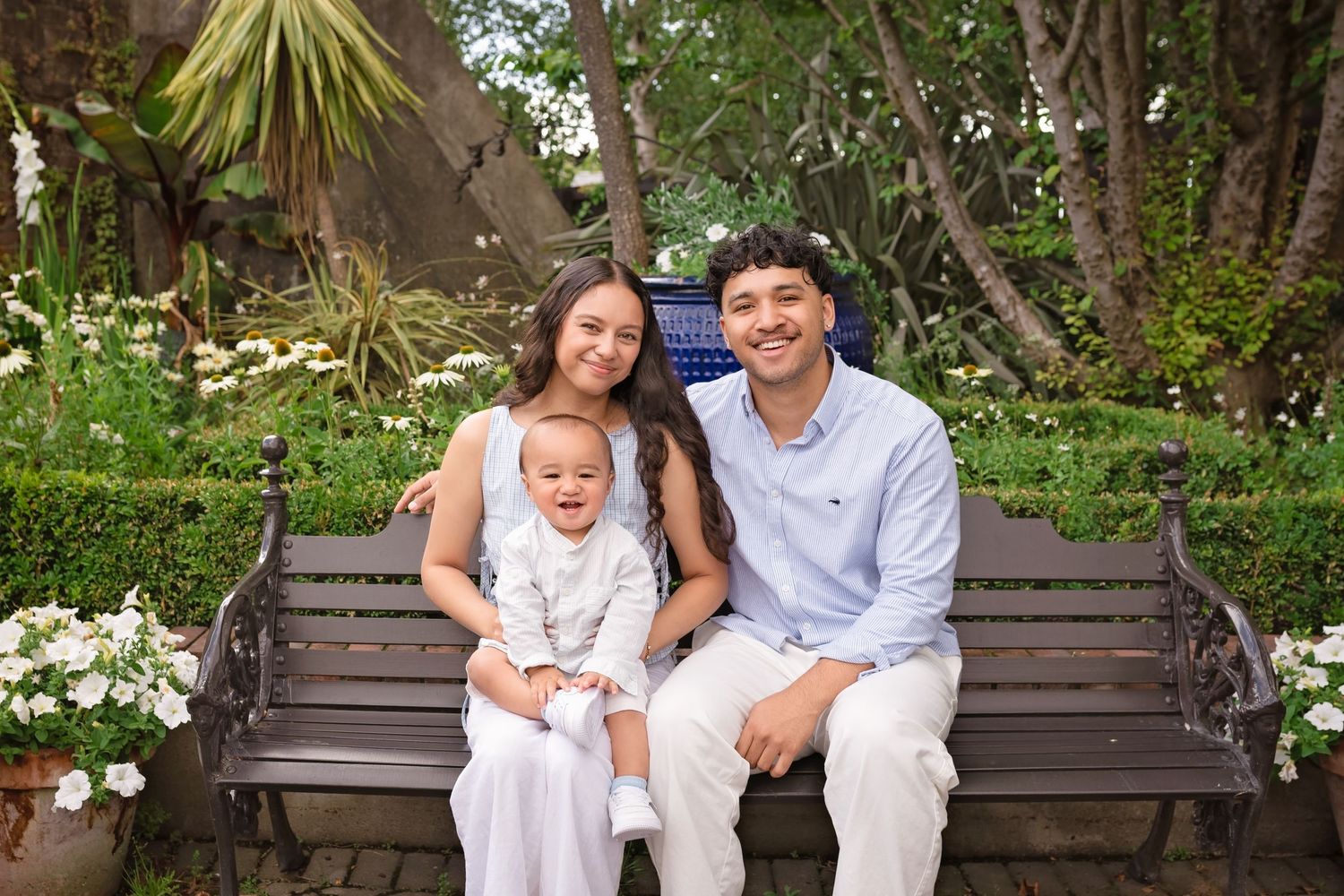Young family with baby sitting on a garden bench surrounded by white flowers and lush greenery.