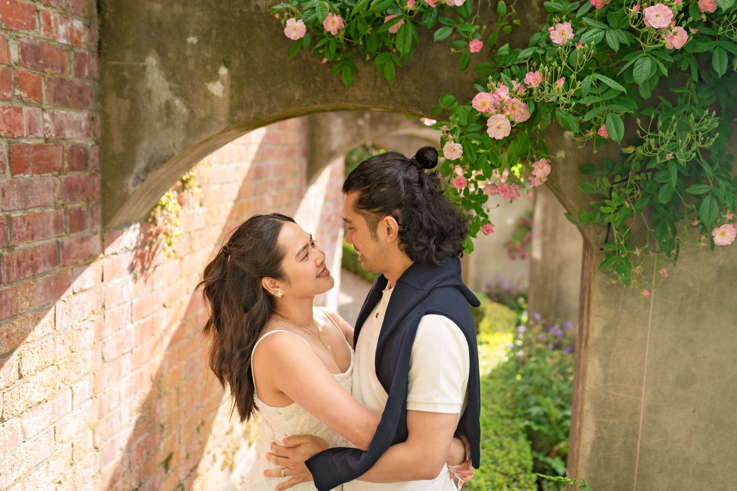 Romantic couple embracing under a rose-covered stone arch, surrounded by lush greenery and pink blooms.