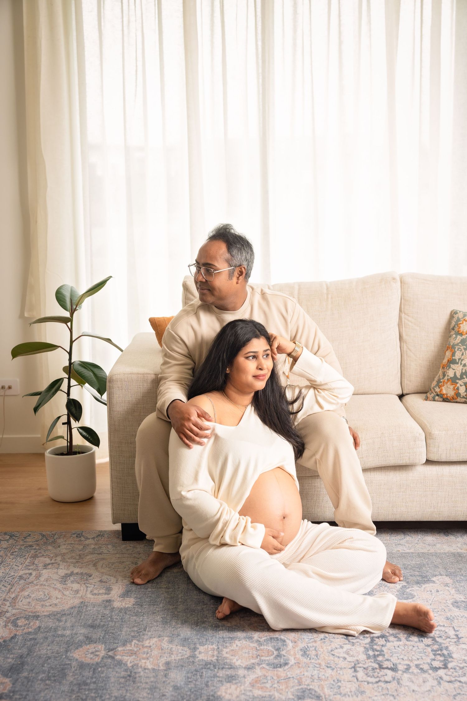 Couple in white loungewear sits together on a rug in front of a couch with a potted plant nearby in serene home setting.