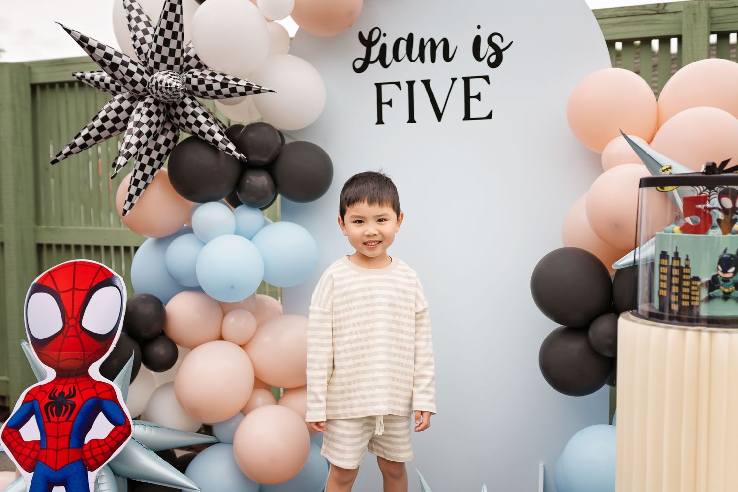 A young boy stands in front of a balloon arch backdrop celebrating Liam turning five, with a Spider-Man balloon nearby.