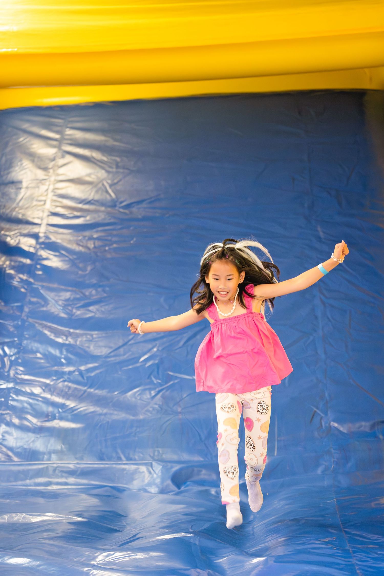 Young girl in pink dress joyfully jumping in a blue inflatable bounce house.