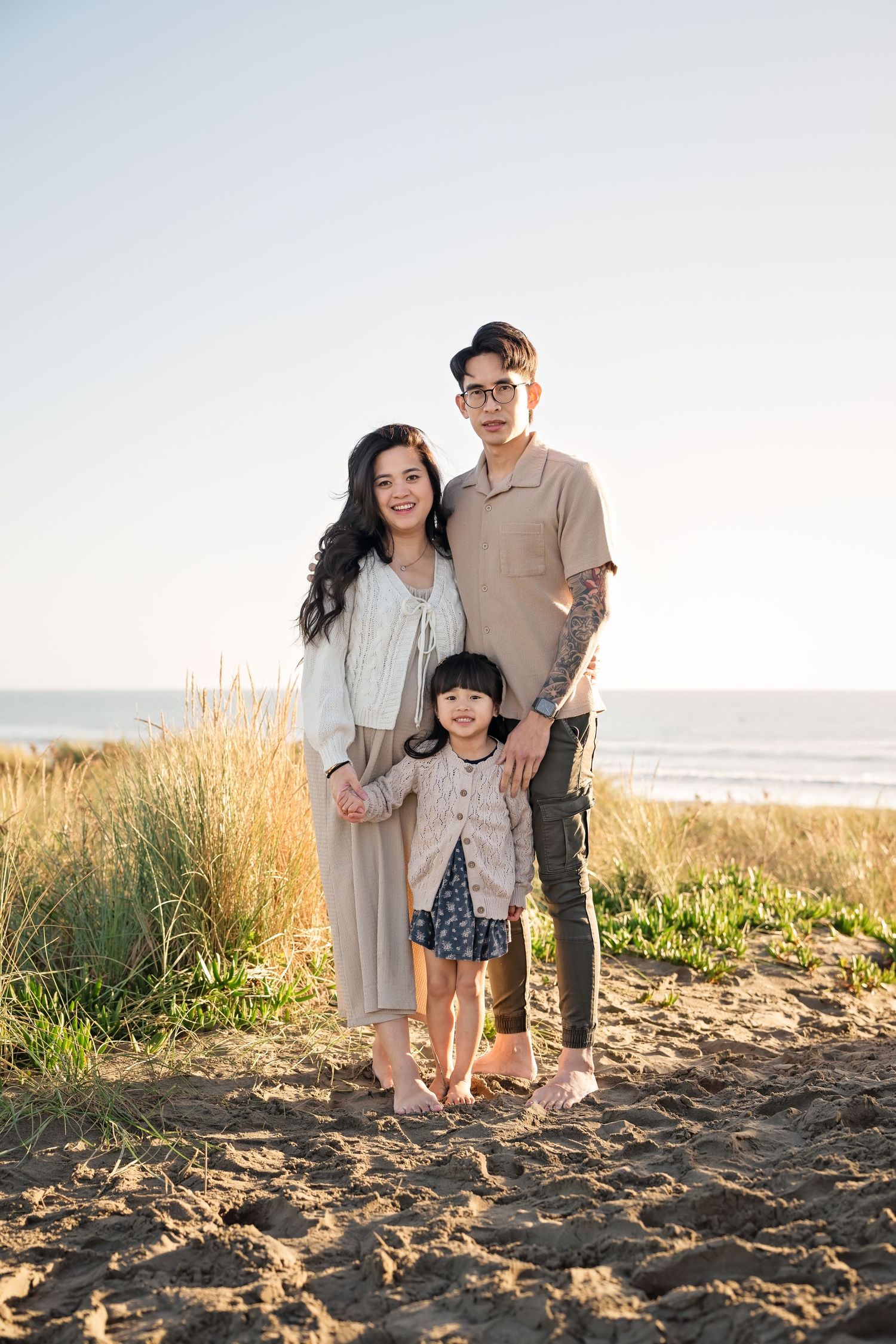 Young Asian family of three posing on sandy beach, parents smiling with young daughter, ocean and grass in background.