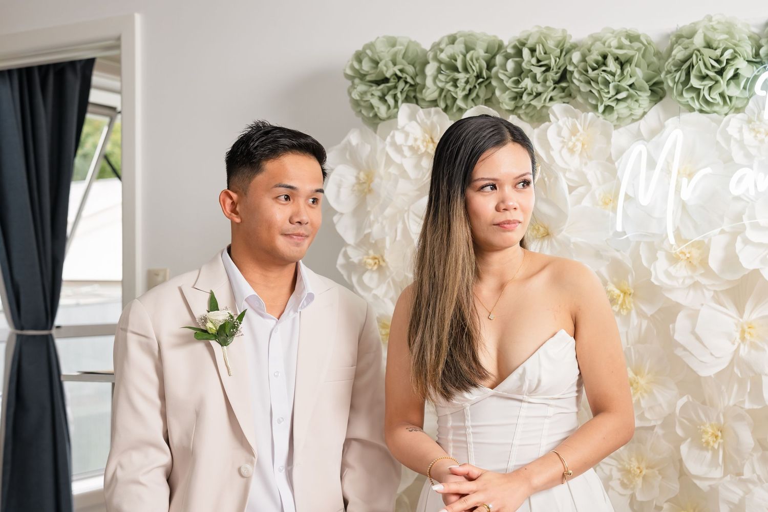 Asian couple in wedding attire pose together in front of white and green floral backdrop.