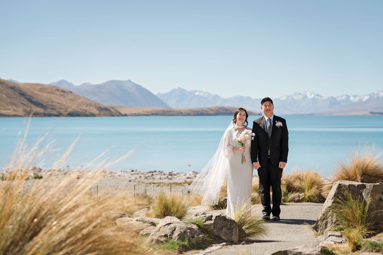 Bride and groom pose together on scenic lakeside path with turquoise water and snowy mountains behind.
