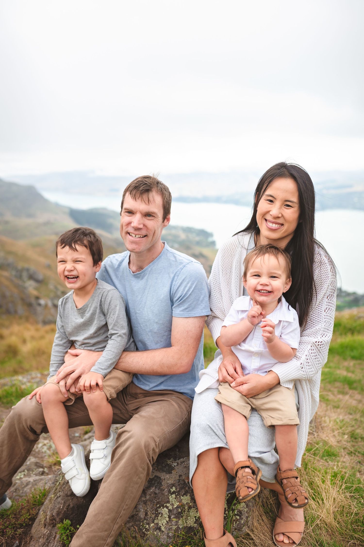 Happy family of four sitting together on rocky outcrop with scenic mountain vista behind them.